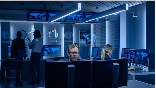 A dark, modern security operations center (SOC) filled with large screens displaying real-time threat analysis and network monitoring data. Several analysts are seen working, with lines of code and network diagrams visible on the screens.