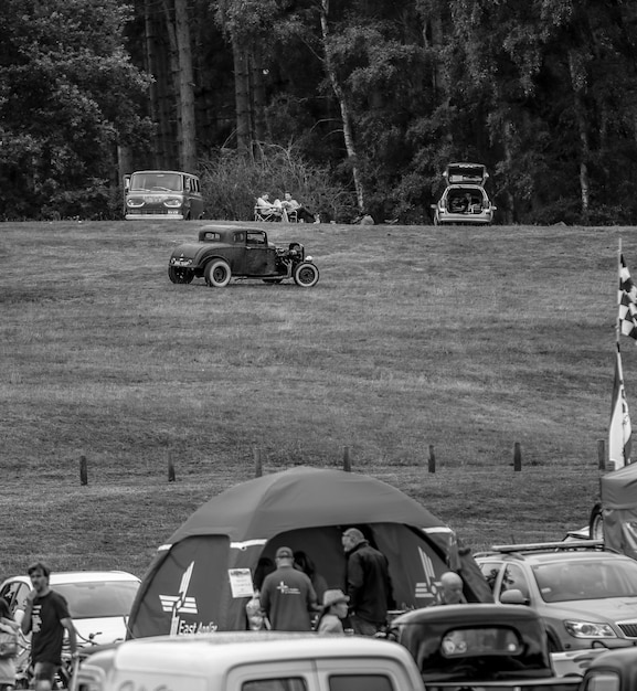 A vintage black and white photograph of a car show from the 1950s, showing classic cars lined up on a grassy field, with families picnicking nearby and admiring the vehicles.