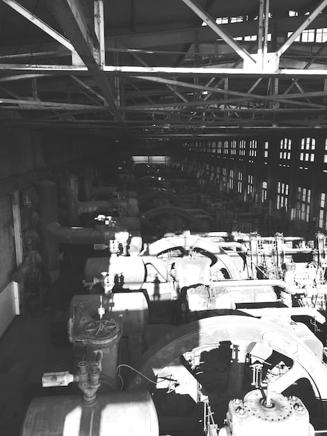 A black and white photograph depicting a bustling automotive repair shop in the 1930s. Mechanics in overalls are working on various vintage cars, with tools and spare parts scattered around the shop floor. The scene captures the early days of automotive repair and restoration, showcasing the hands-on craftsmanship and dedication of the era. The shop is filled with vintage signs and advertisements, adding to the historical atmosphere and highlighting the evolution of car care and restoration practices.