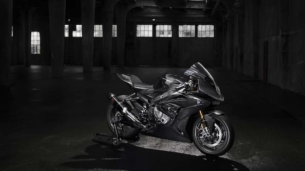 A profile view of the Triumph Speed Triple 1200 RS, highlighting its aggressive stance and muscular design. The motorcycle is parked on a racetrack, emphasizing its sporty capabilities. The background includes other high-performance motorcycles and pit lane equipment.