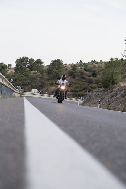 The 2025 Suzuki DR650S riding on a paved road, showcasing its upright riding position and basic tires. The scenery includes a winding road through a forest, with the focus on the bike's on-road handling.